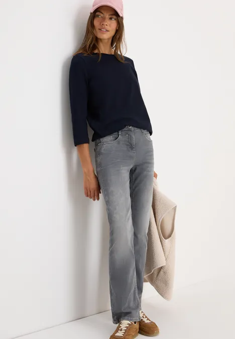 Frau mit dunkelblauem Pulli, grauer Jeans und rosa Kappe lehnt an einer weißen Wand.